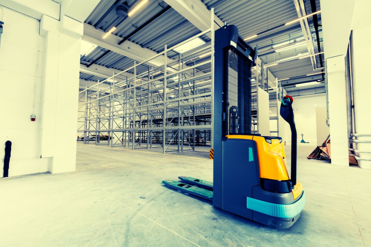how-high-does-a-pallet-jack-lift1 Image of an electric pallet jack in a warehouse with tall shelving racks, illustrating how high does a pallet jack lift for heavy-duty operations.