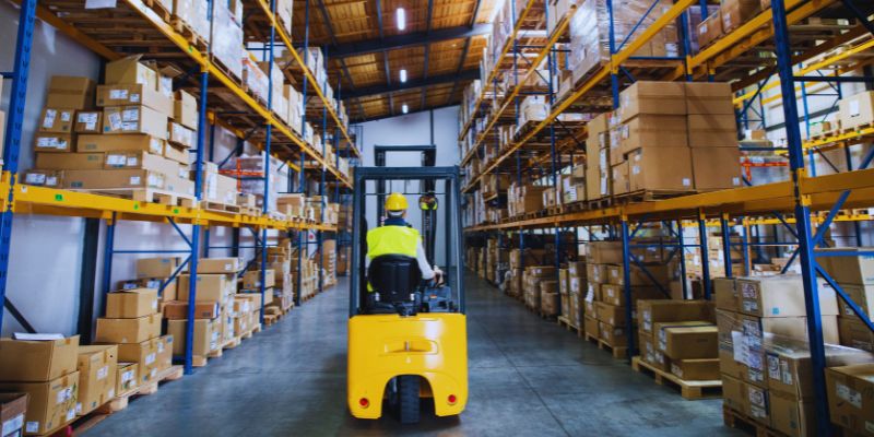 Forklift driver in high-visibility vest operating a yellow electric forklift between tall racking aisles, essential item on every comprehensive warehouse equipment list.