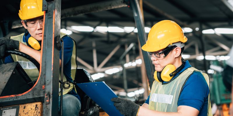 forklift-classifications1 Two safety-equipped workers in hard hats and high-visibility vests reviewing a forklift's controls and documentation inside an industrial facility, illustrating different forklift classifications and safety standards.
